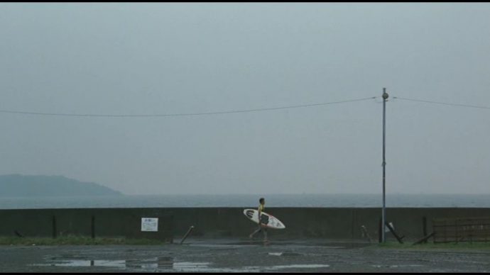 Shigaru caminha com roupa de surf e uma prancha em meio a chuva.
