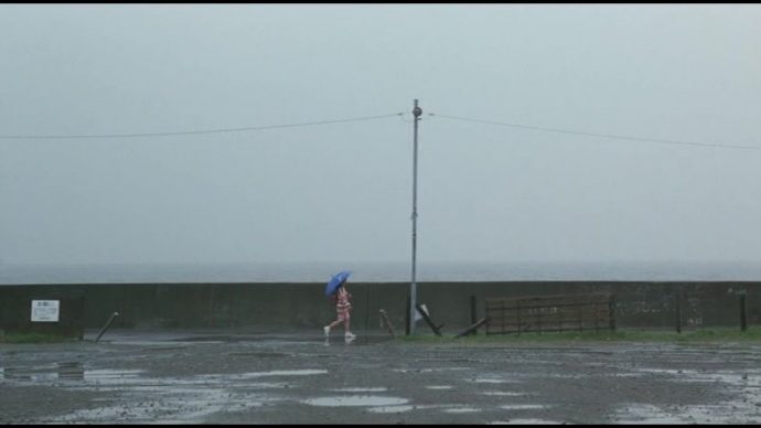 Takako caminha em meio a chuva om um guarda-chuva azul. 