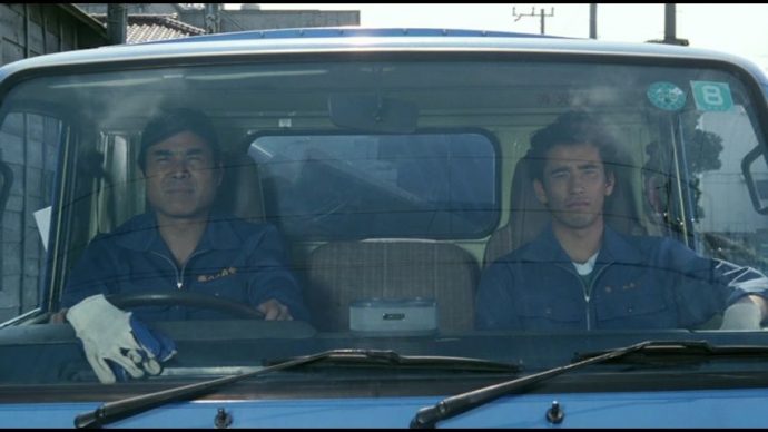 Shigaru, o protagonista de "O Mar Mais Silencioso Daquele Verão", e outro homem olham pela janela do caminhão de um caminhão de lixo. Os dois usam uniforme de garis.