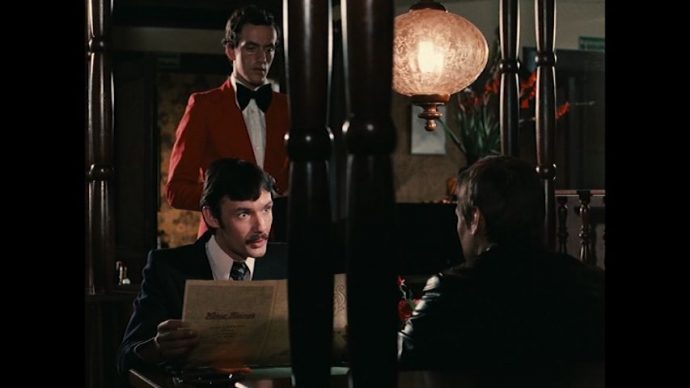 Fox e Eugen estão em um restaurante chique, entre ele há no primeiro plano uma estrutra de madeira que corta o plano ao meio.