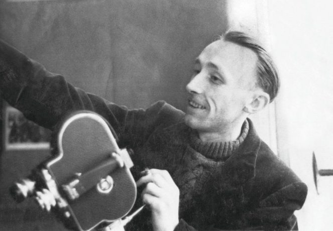 Foto do de um dos críticos mais importantes da história do cinema, o francês André Bazin. Na imagem o crítico sorri, segura uma câmera portátil e tem o braço esticado para fora do enquadramento.