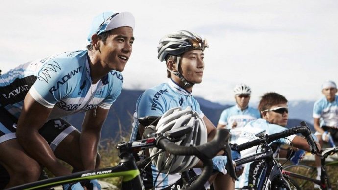 Eddie Peng e Jeong Ji-Won, em cena do filme To the Fore. Nela, os dois estão montados em uma bicicleta e usam roupas azuis de uma equipe de ciclismo. Ao fundo mais três membros da equipe. AO longe ainda se pode ver, meio desfocada, uma montanha, eles parecem estar parados em um lugar alto, como uma serra ou a subida de uma colina