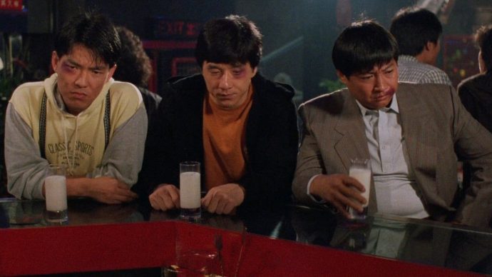 Yuen Biao, Jackie Chan e Sammo Hung em uma cena de algum filme de Hong Kong. Os três parecem ter levado uma surra, estão sentados diante de um balcão de um bar, cada um com um copo de leite diante de si. Os três homens têm uma expressão de chateação e cansaço no rosto. 
