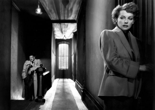 Um corredor de um prédio. Em primeiro plano a atriz Ann Sheridan está encostada em uma porta como se estivesse receosa de entrar. Ela observa de canto de olho o personagem de Robert Keith que a observa da escada, ele está apoiado no corrimão em pose de espera. 